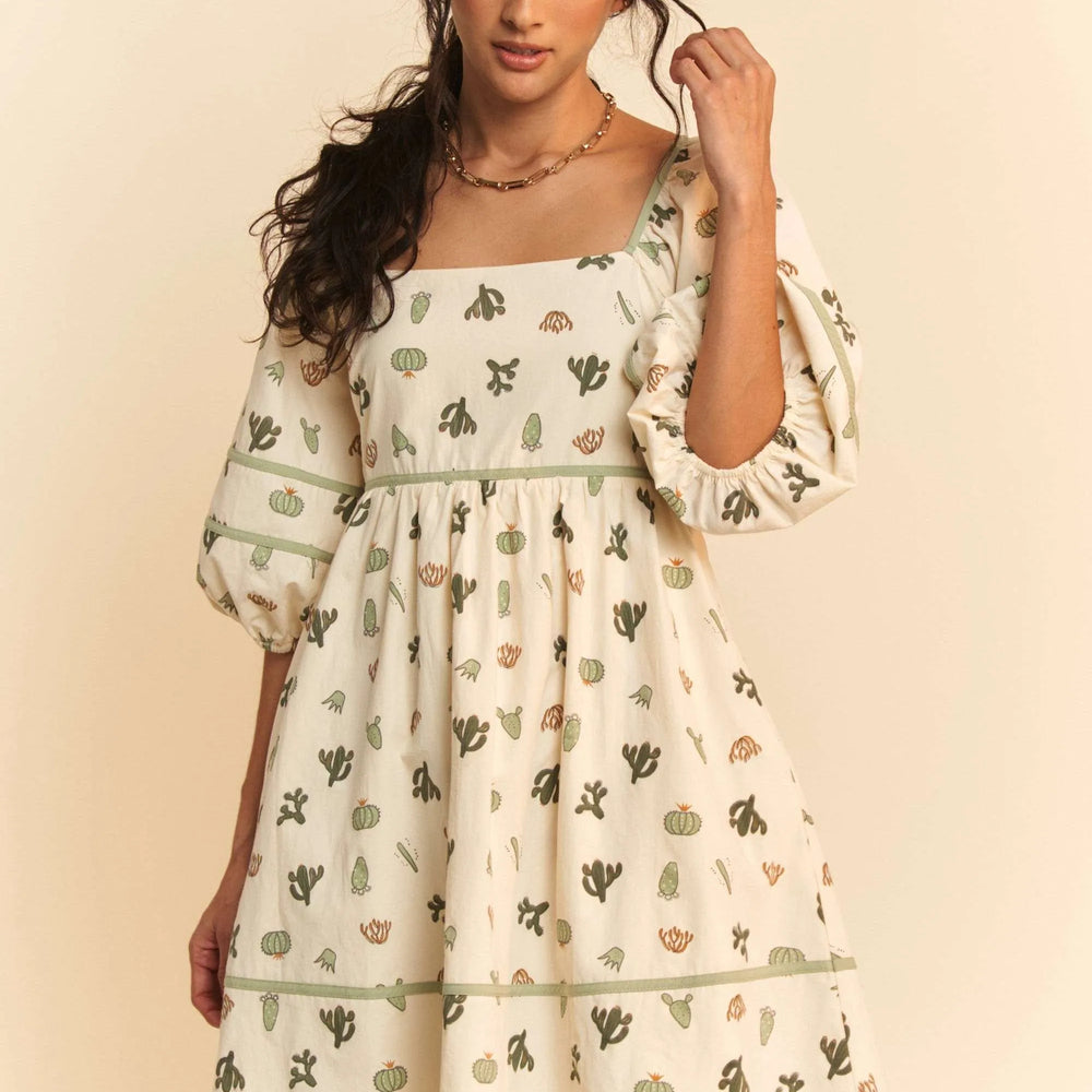 Model wearing a cactus print poplin mini dress from Unique Kulture, featuring contrast piping, smocked bodice, and tied straps.

 close front view