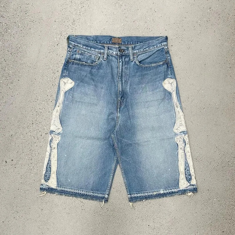 Blue denim shorts with white skeleton bone patches on each side and raw frayed hems, laid flat on a concrete background.



