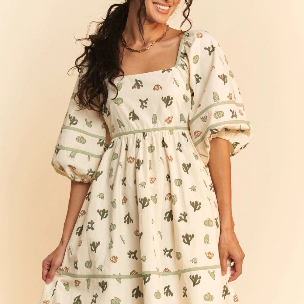 Model wearing a cactus print poplin mini dress from Unique Kulture, featuring contrast piping, smocked bodice, and tied straps.

