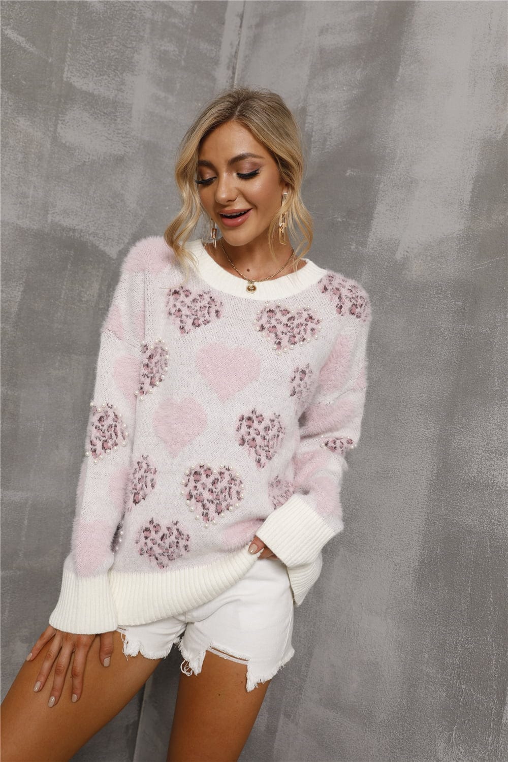 Heart Pattern Round Neck Long Sleeve Sweater by Unique Kulture. The sweater features a cozy round neckline and long sleeves. The pattern on the sweater consists of intricate heart shapes arranged in a repetitive and stylish design, adding a touch of romance to the overall look. The color palette includes soft shades of pink, red, and white, creating a harmonious and eye-catching contrast