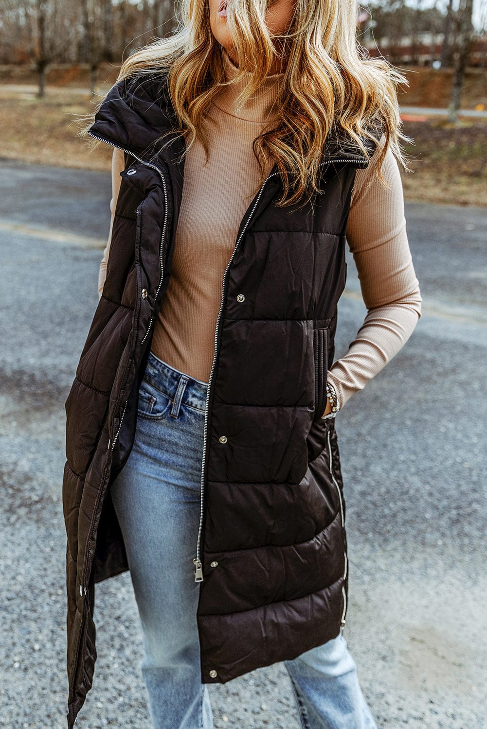 "Image: A model wearing the Unique Kulture Longline Hooded Sleeveless Puffer Vest. The vest is a deep black color with a sleek, quilted design. It features a longline cut, extending below the hips, and has a hood attached. The front of the vest has a zipper closure, and there are two zippered pockets on the sides. The model is standing outdoors, and the vest adds a stylish and warm layer to their outfit."