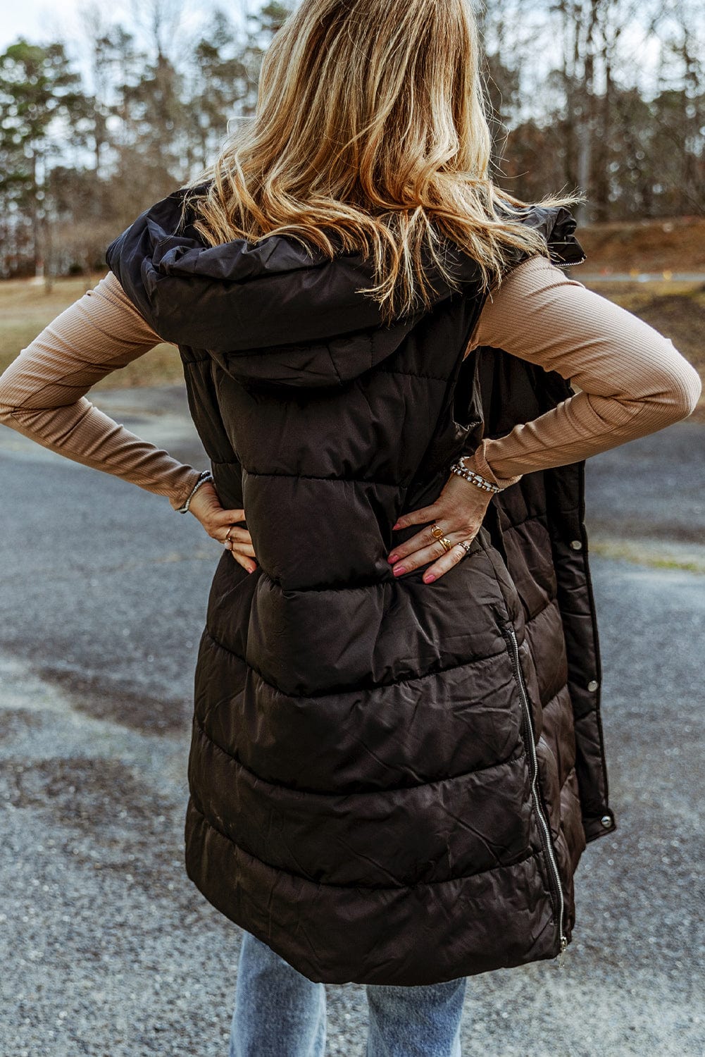 "Image: A model wearing the Unique Kulture Longline Hooded Sleeveless Puffer Vest. The vest is a deep black color with a sleek, quilted design. It features a longline cut, extending below the hips, and has a hood attached. The front of the vest has a zipper closure, and there are two zippered pockets on the sides. The model is standing outdoors, and the vest adds a stylish and warm layer to their outfit."